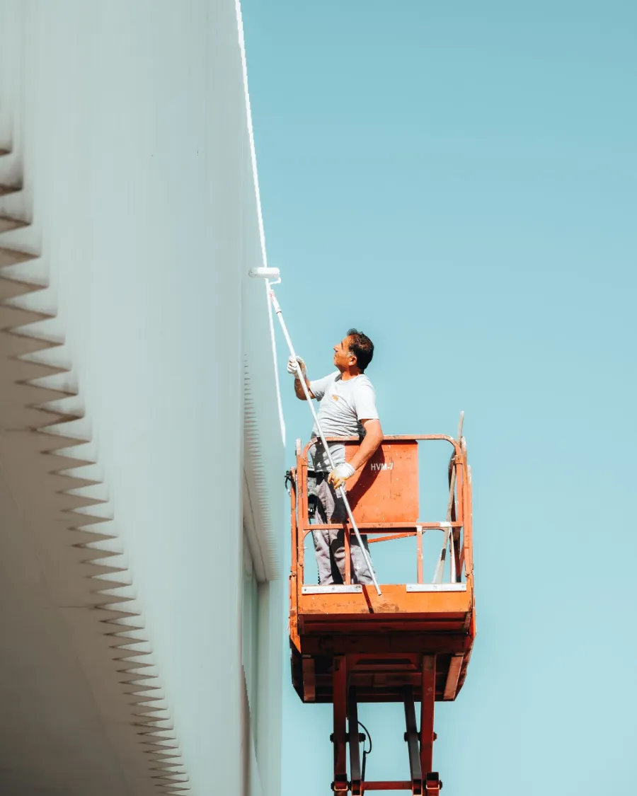 Pintor em plataforma elevatória aplicando tinta na fachada de um prédio, contra céu limpo.